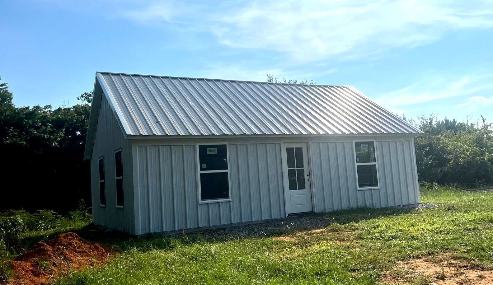Small, light blue-sided cabin with metal roof, windows, and door, on grassy field under a blue sky.