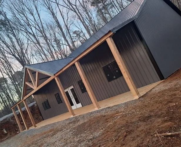 Dark gray barn with wooden porch and exposed rafters in a wooded area.