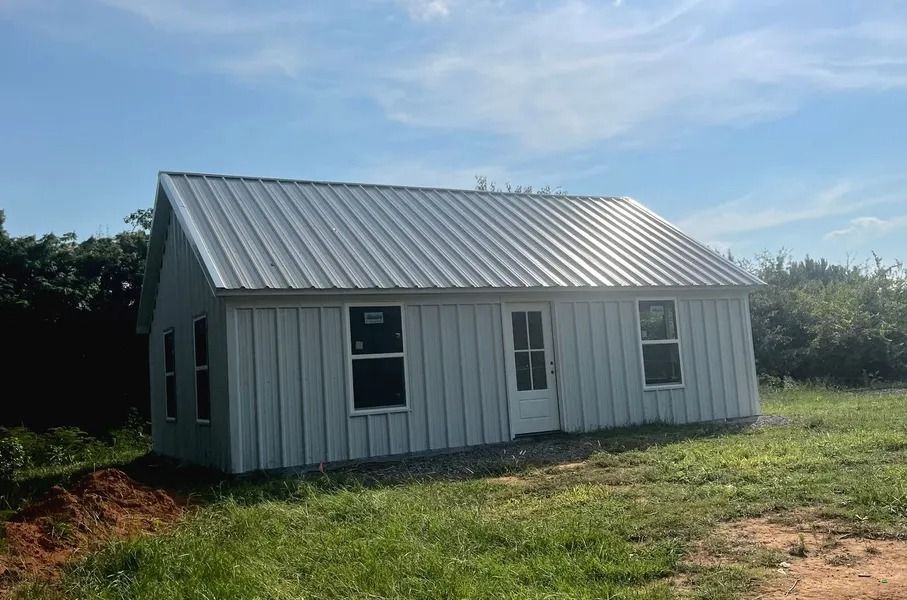 Small white-sided building with a metal roof in a grassy field under a blue sky.