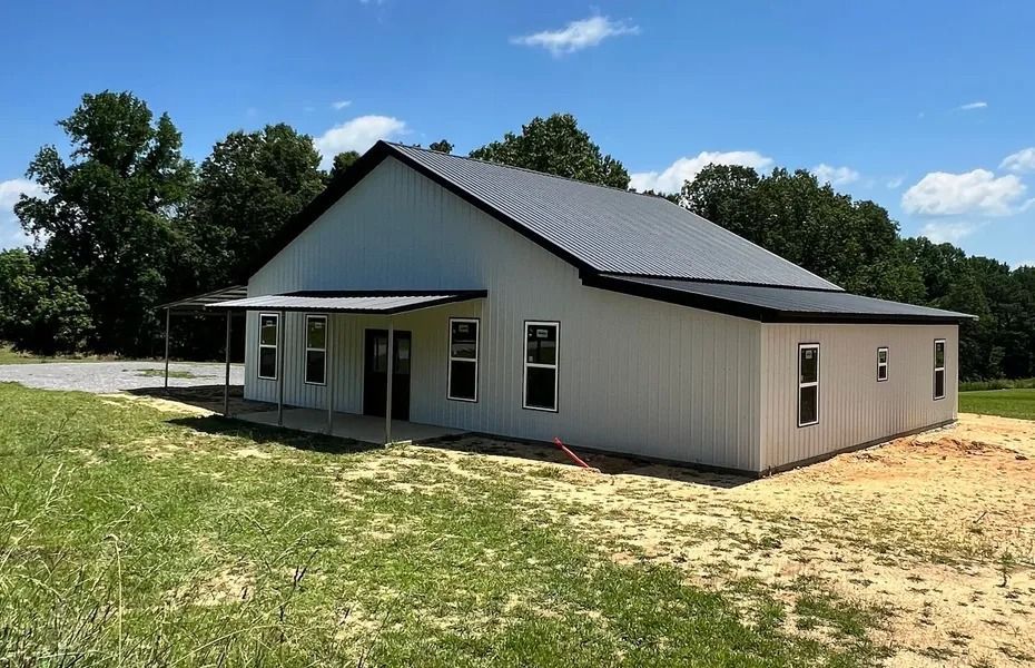 White metal barn-style building with black roof and porch, set in a grassy field under a blue sky.