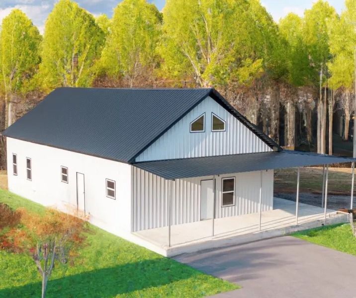 White, barn-style house with black roof and covered porch in a grassy setting with trees.