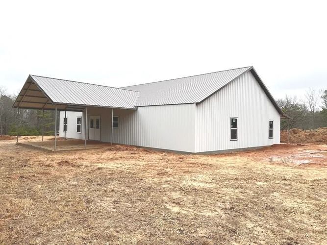 White metal-sided building with a carport; set in a field with some trees.