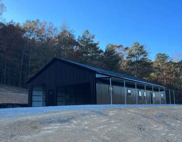 Black metal barn with open garage door, surrounded by trees and gravel driveway.