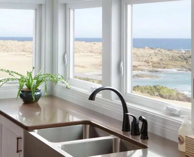 Kitchen with windows overlooking a beach; a sink, faucet, and plant sit on the countertop.
