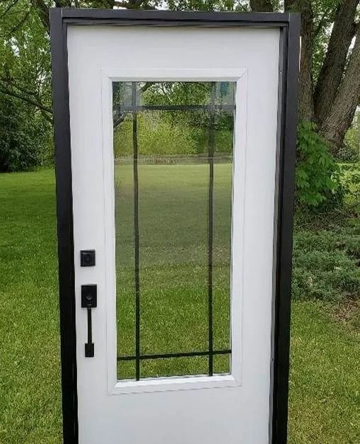 White door with black trim, a large glass window, and black hardware, set in a grassy outdoor space.