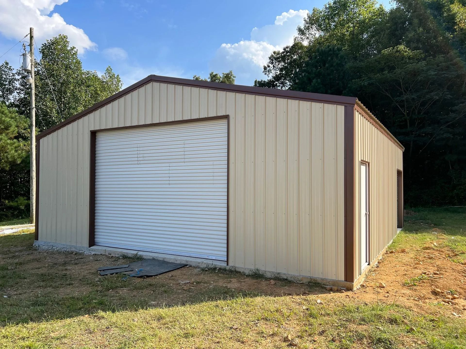 Tan metal garage with brown trim and closed garage door; set in a grassy yard with trees in the background.