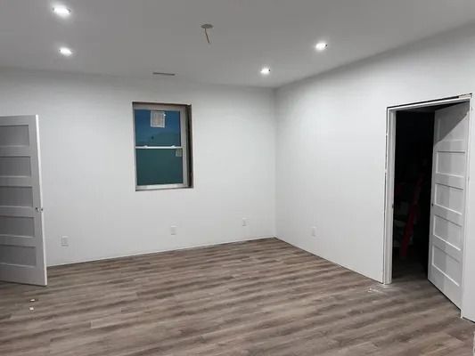 Empty room with white walls, brown floor, window, and two doorways. Recessed lighting.
