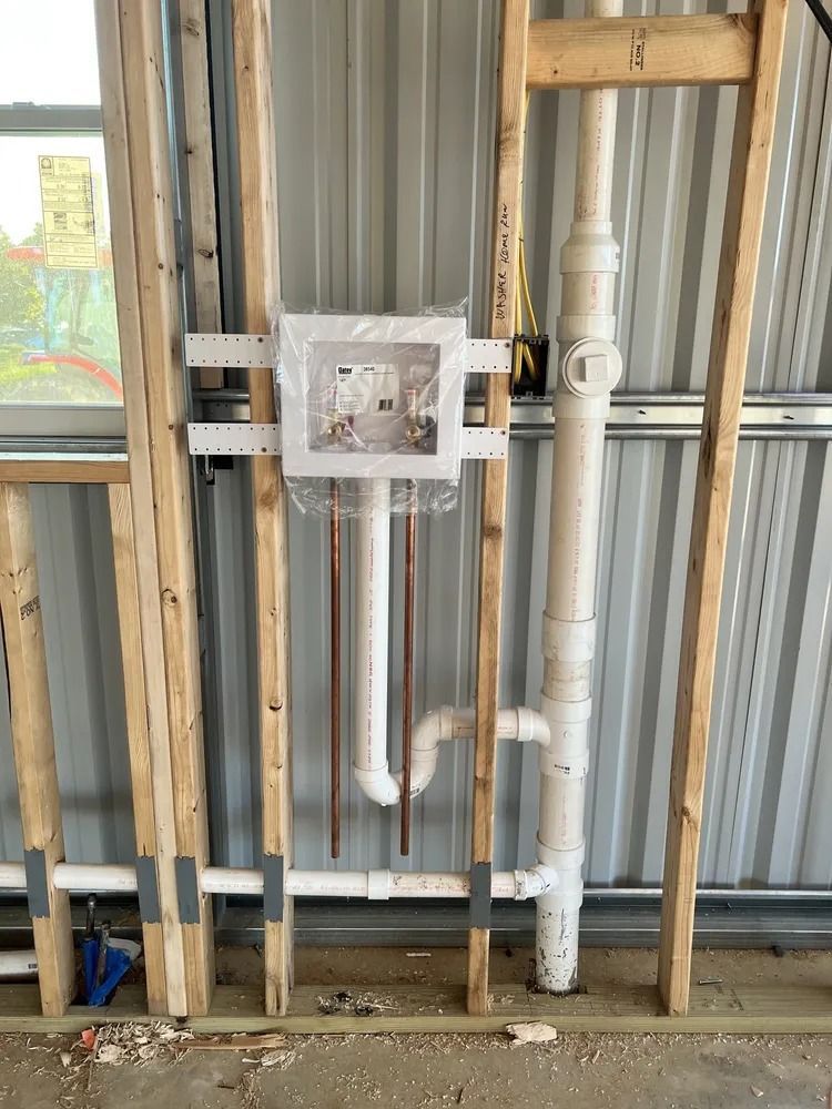Plumbing rough-in: Copper pipes for water, white drain pipes within a wooden wall frame, near a window.