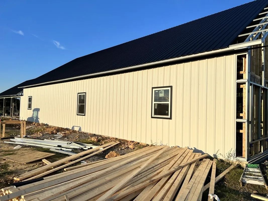 Exterior of a building under construction. Cream siding, black roof, windows, lumber piles, blue sky.