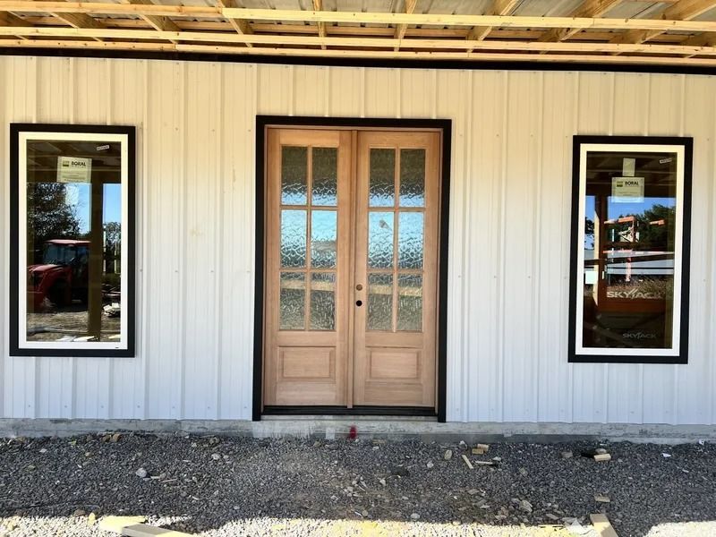 Light-colored wooden double doors with clear panes, flanked by two vertical windows. Building exterior under construction.