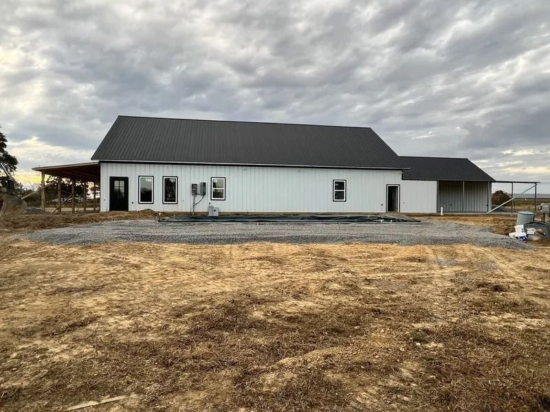 White farmhouse with black roof, dark windows and door, gravel yard, cloudy sky.
