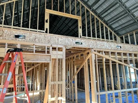 Interior of a wooden house under construction, showing framing, window, and a ladder.