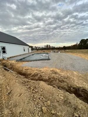 Construction site with trench, gravel, and a partially-covered pool next to a white house under cloudy skies.