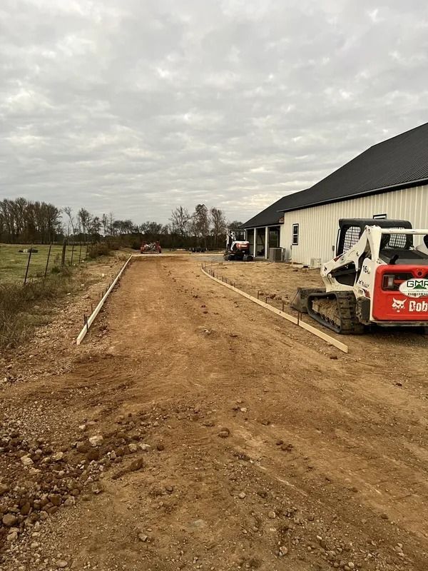 Dirt driveway under construction next to a white building; small tractor present. Overcast sky.