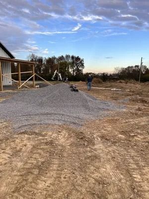 Pile of gravel next to a partially built house; a person stands nearby in a field under a blue sky.