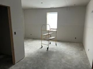 A room under construction with bare concrete floor, drywall walls, and a wheeled cart near the window.
