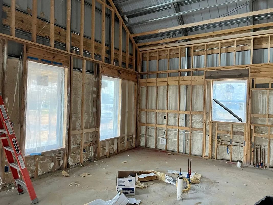 Interior of a building under construction, framed walls, windows, insulation, and a ladder are visible.