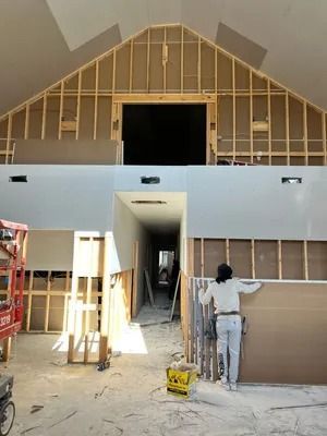 Construction site: A person installing drywall. Wood framing, long hallway, and an open gable roof are visible.