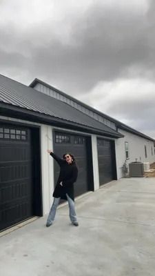 Person in black coat and jeans poses in front of a garage with dark doors on a cloudy day.
