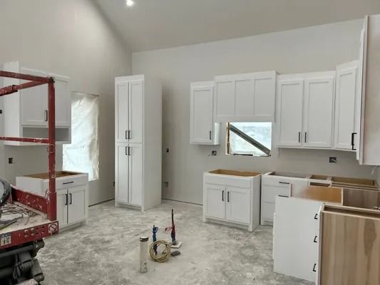 White kitchen cabinets in a room under construction. Tall cabinet, upper and lower cabinets, unfinished floor, and lift.