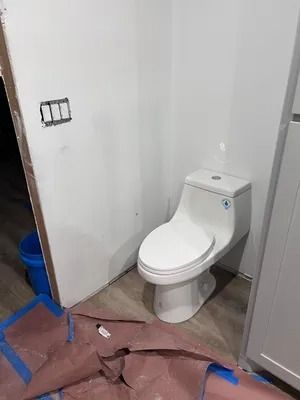 White toilet in a small bathroom corner, surrounded by white walls, with some construction materials on the floor.