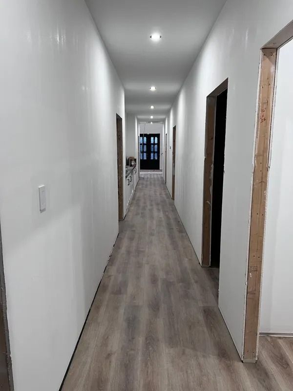 Long, white hallway with wooden floor and doorways, leading to a dark door at the end.