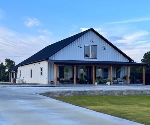 White barn-style house with black roof, covered porch, and concrete driveway against a blue sky.