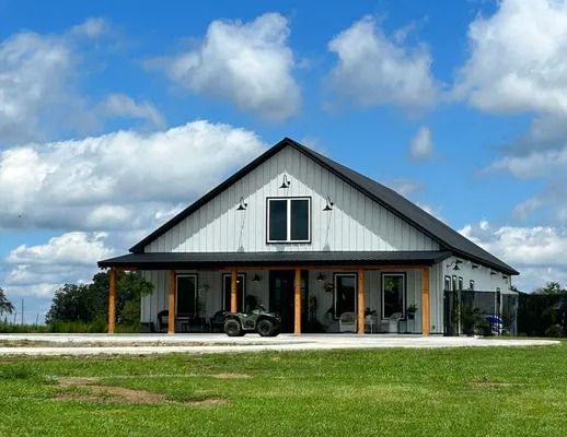 White farmhouse with black roof and porch, set in green field under blue sky.