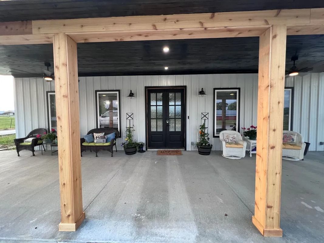 Covered porch with wooden beams, white siding, black door, and seating.