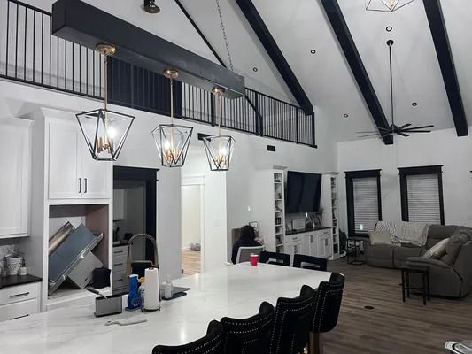 Kitchen with high ceiling, black beams, white cabinets, island with chairs, and hanging lights.