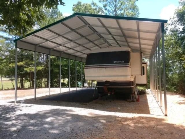 RV parked under a metal carport with a green roof, in a gravel driveway, trees in the background.