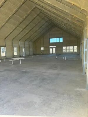 Interior of a large, unfinished building with high, vaulted ceiling. Concrete floor, beige walls, windows, and doors.