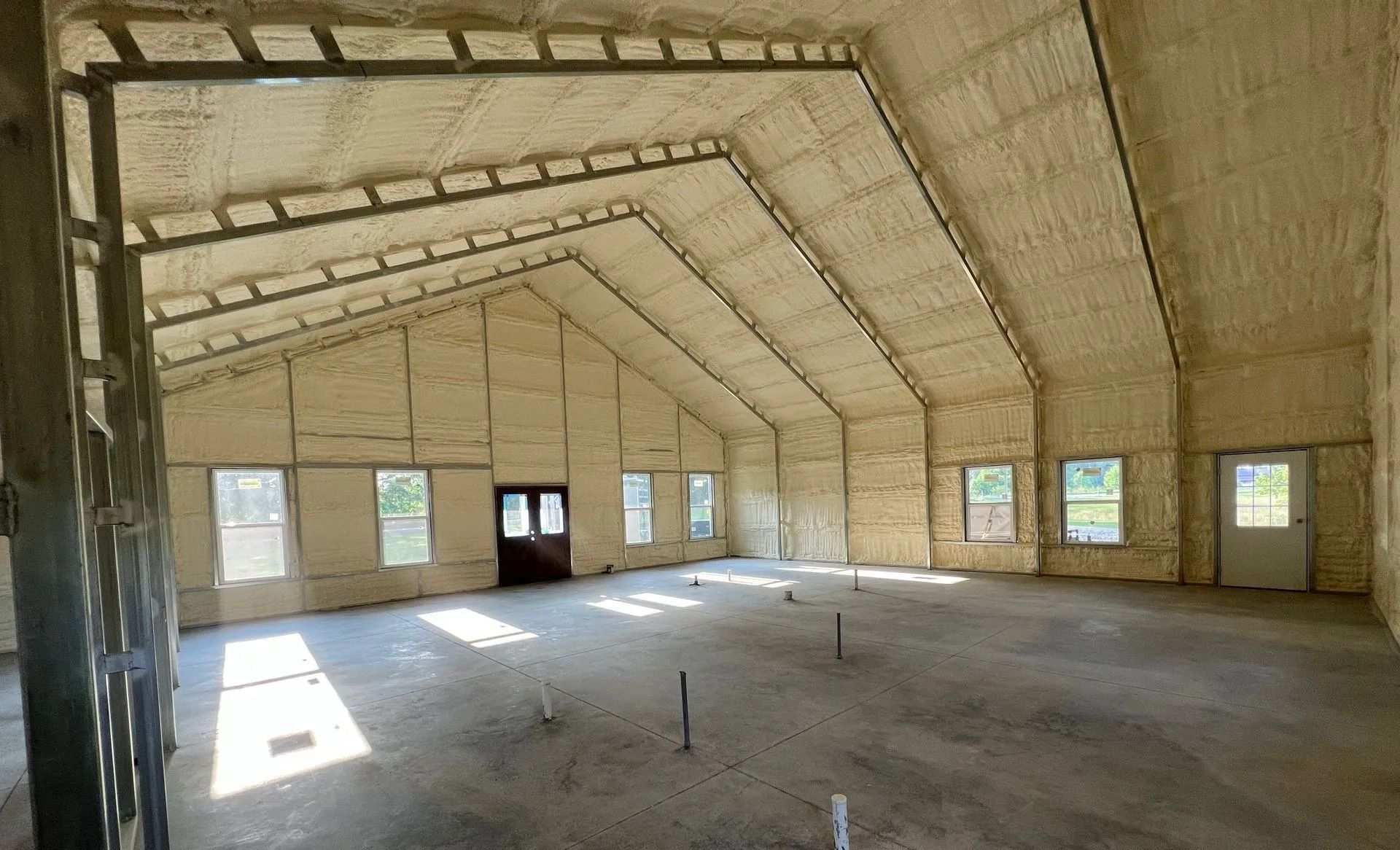 Interior of a large building under construction with foam insulation on walls and angled ceiling. Concrete floor, windows, and door visible.
