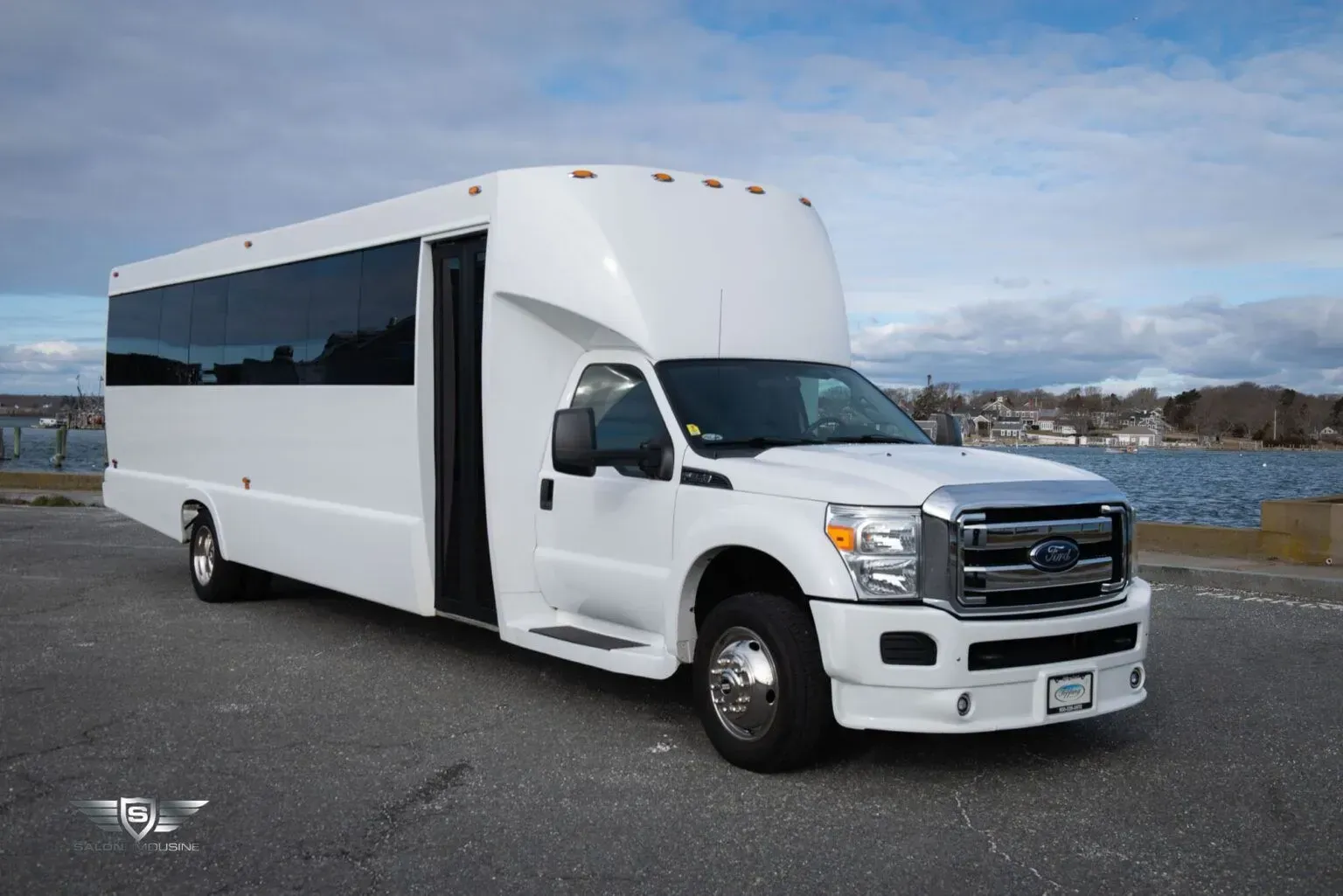 White party bus parked on a paved lot near a body of water with a cloudy sky.