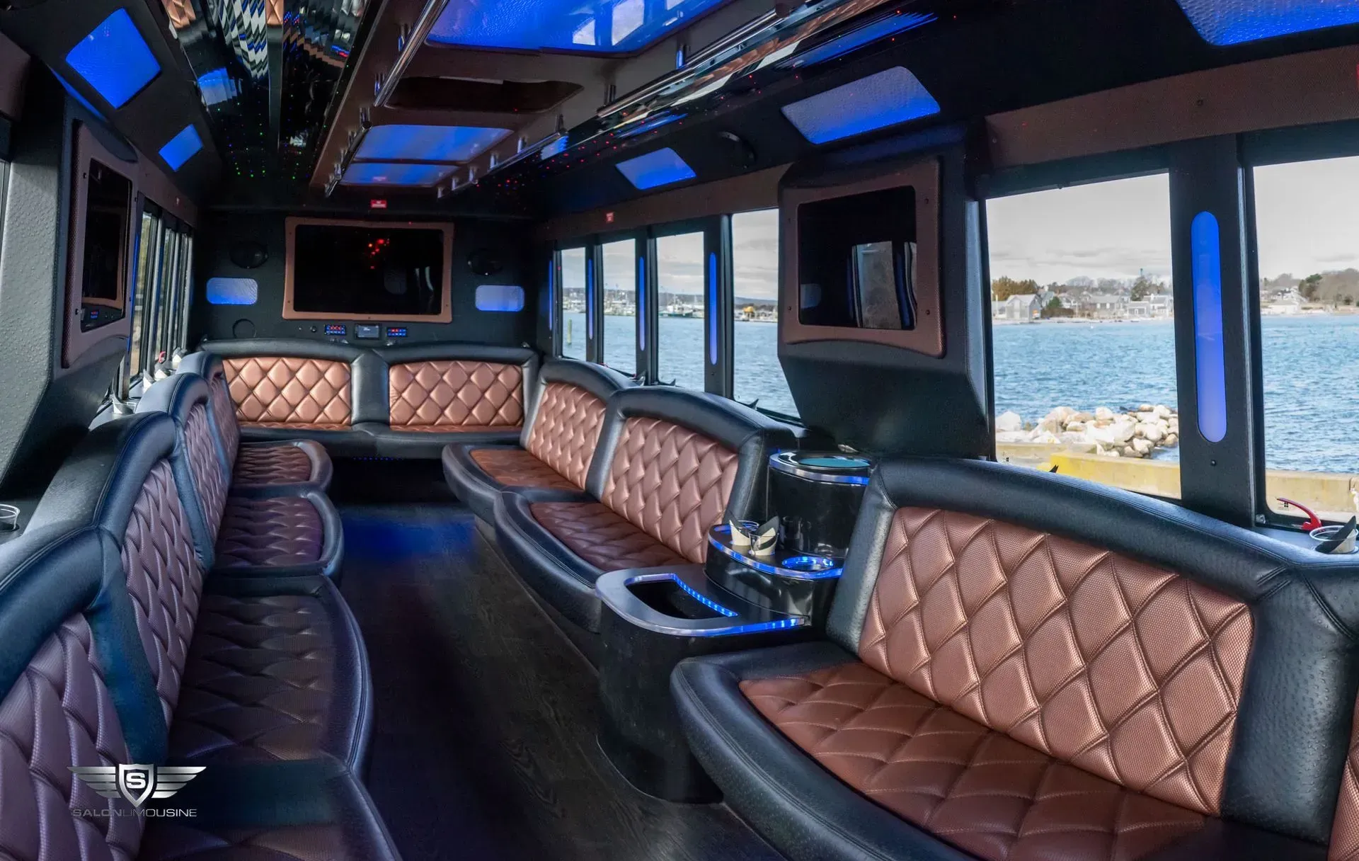 Interior of a luxury party bus with brown leather seating, multiple screens, and blue lighting, overlooking a water view.