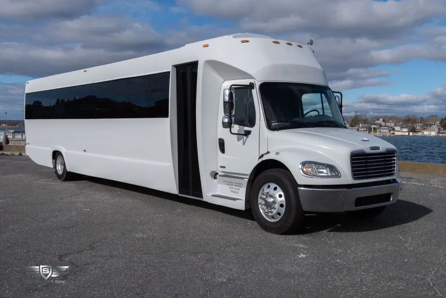 White shuttle bus parked on a concrete surface, with water and buildings in the background. The door is open.