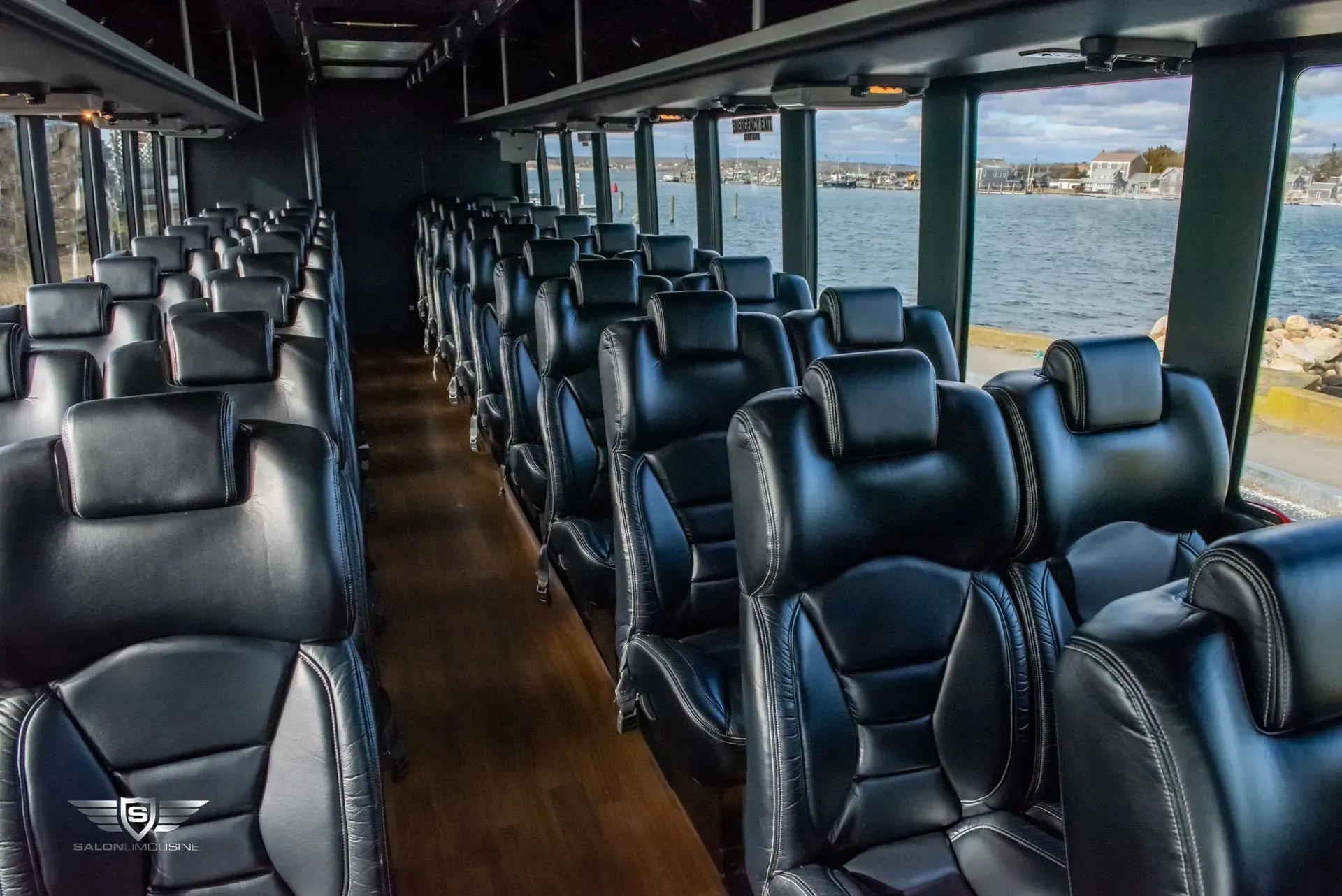 Interior of a bus with rows of black leather seats, a wooden floor, and windows overlooking water.