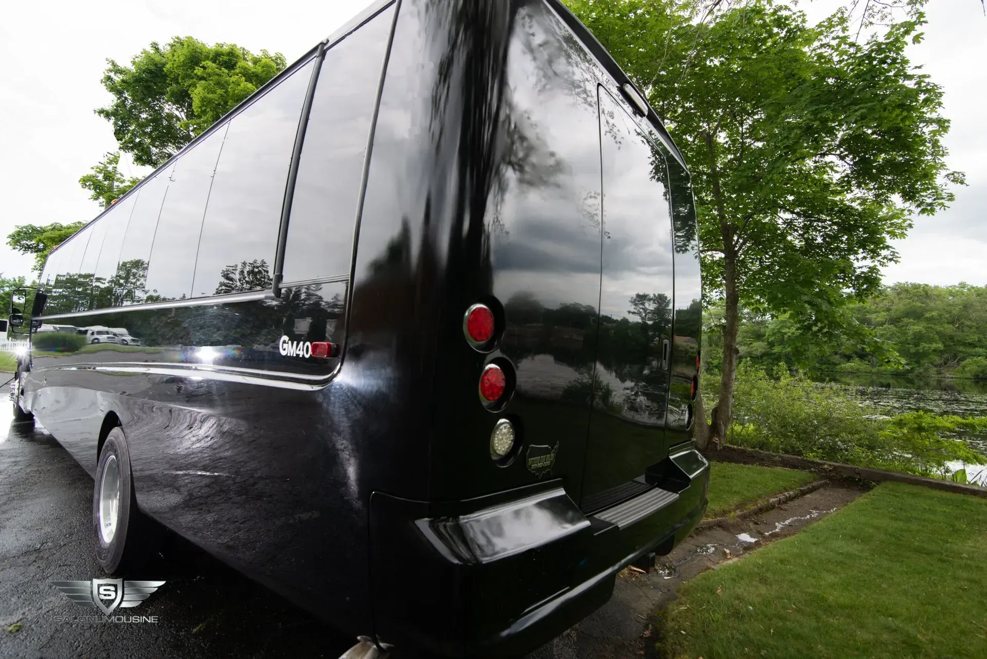 Black party bus parked, reflecting trees and sky. Rear view shows taillights and tinted windows.