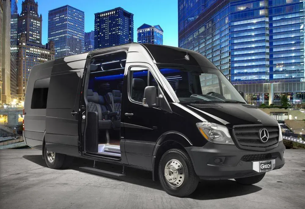 Black Mercedes-Benz passenger van with open sliding door parked in front of a city skyline at night.