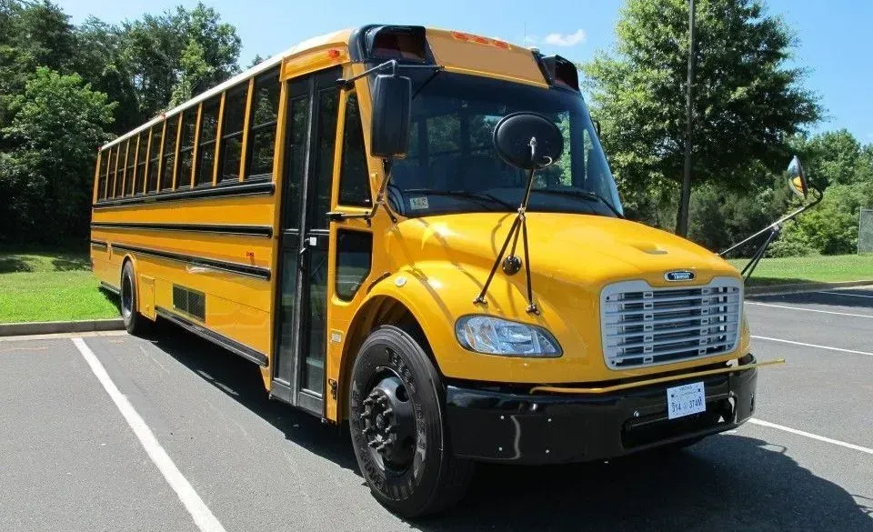 Yellow school bus parked on asphalt, in front of a grassy area, trees visible in background.