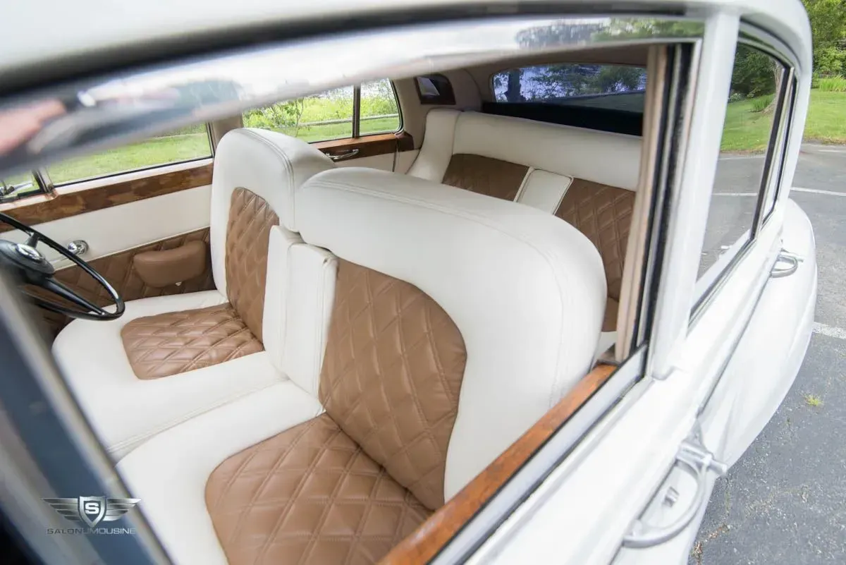Inside view of a white and brown quilted leather interior of a classic car, with wood trim and open door.