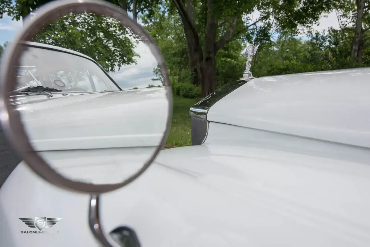 White classic car with hood ornament and side mirror reflecting another car.