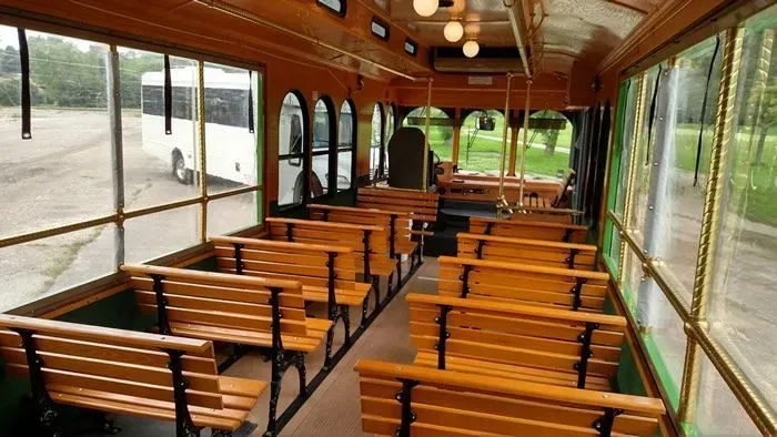 Interior of a trolley with wooden benches, windows, and hanging lights. A white bus is visible outside the window.