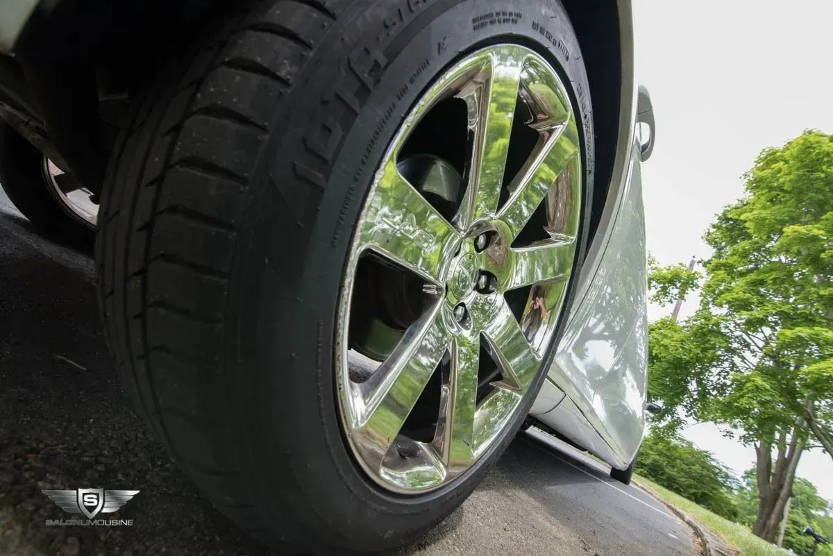 Chrome car wheel on a black tire, parked next to a white car on a paved area, with green foliage.