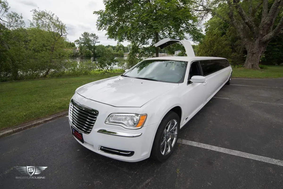 White limousine with gullwing door open, parked on asphalt near water and trees.
