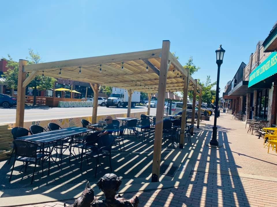 A group of people are sitting under a pergola on a sidewalk.
