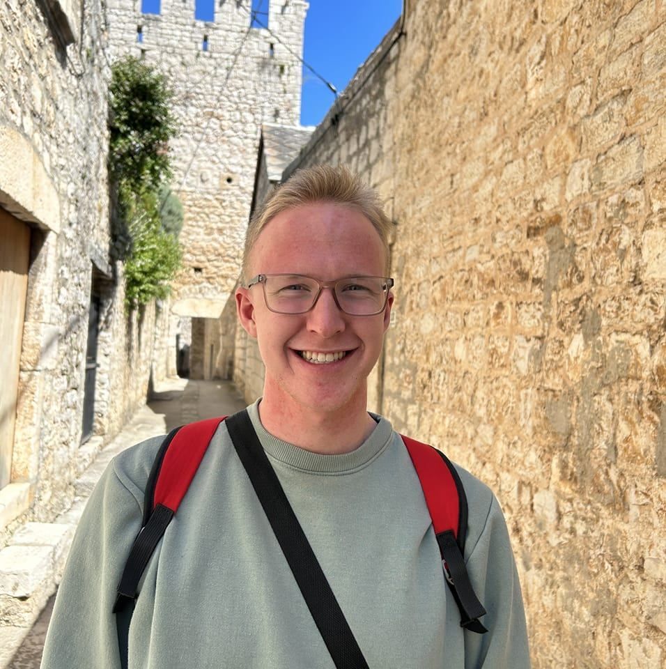 Man with glasses and backpack smiles in a narrow stone alley, with a tower in the background.