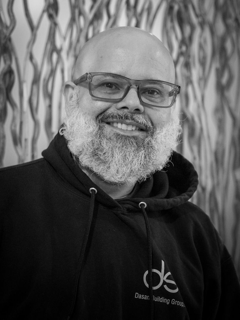 A smiling person with a thick beard and glasses wears a black hooded sweatshirt against a patterned, neutral background.