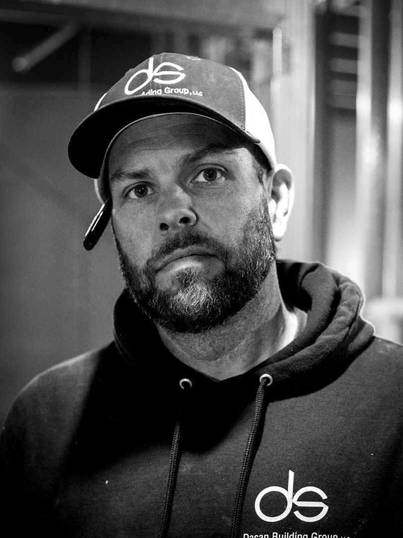 A person with a beard wearing a branded baseball cap and hoodie, looking directly at the camera in a black and white portrait.