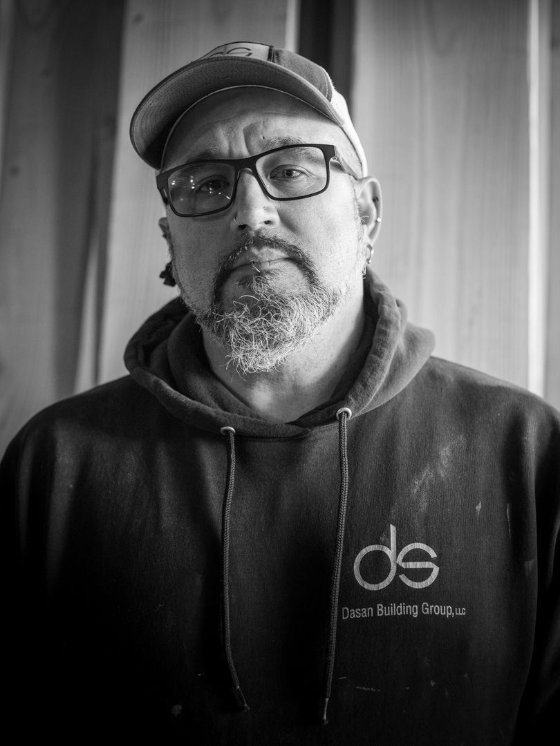 A person with a beard and glasses wears a baseball cap and a dark hooded sweatshirt, centered against a wood-paneled wall.